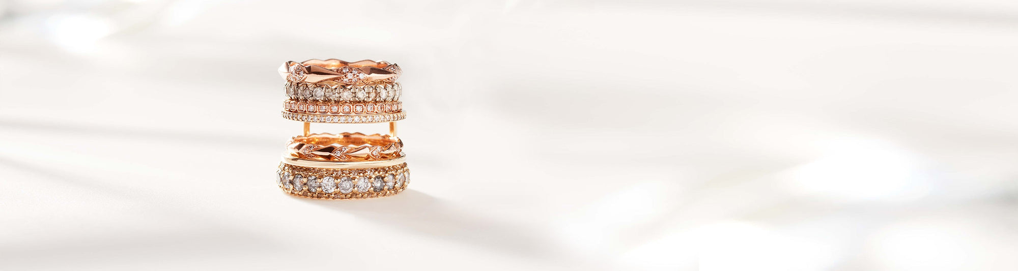 Multiple straight bands stacked on top of each other set in yellow and rose gold. Two bands have white diamond accent stones and two have grey diamond accent stones on a grey and white background.