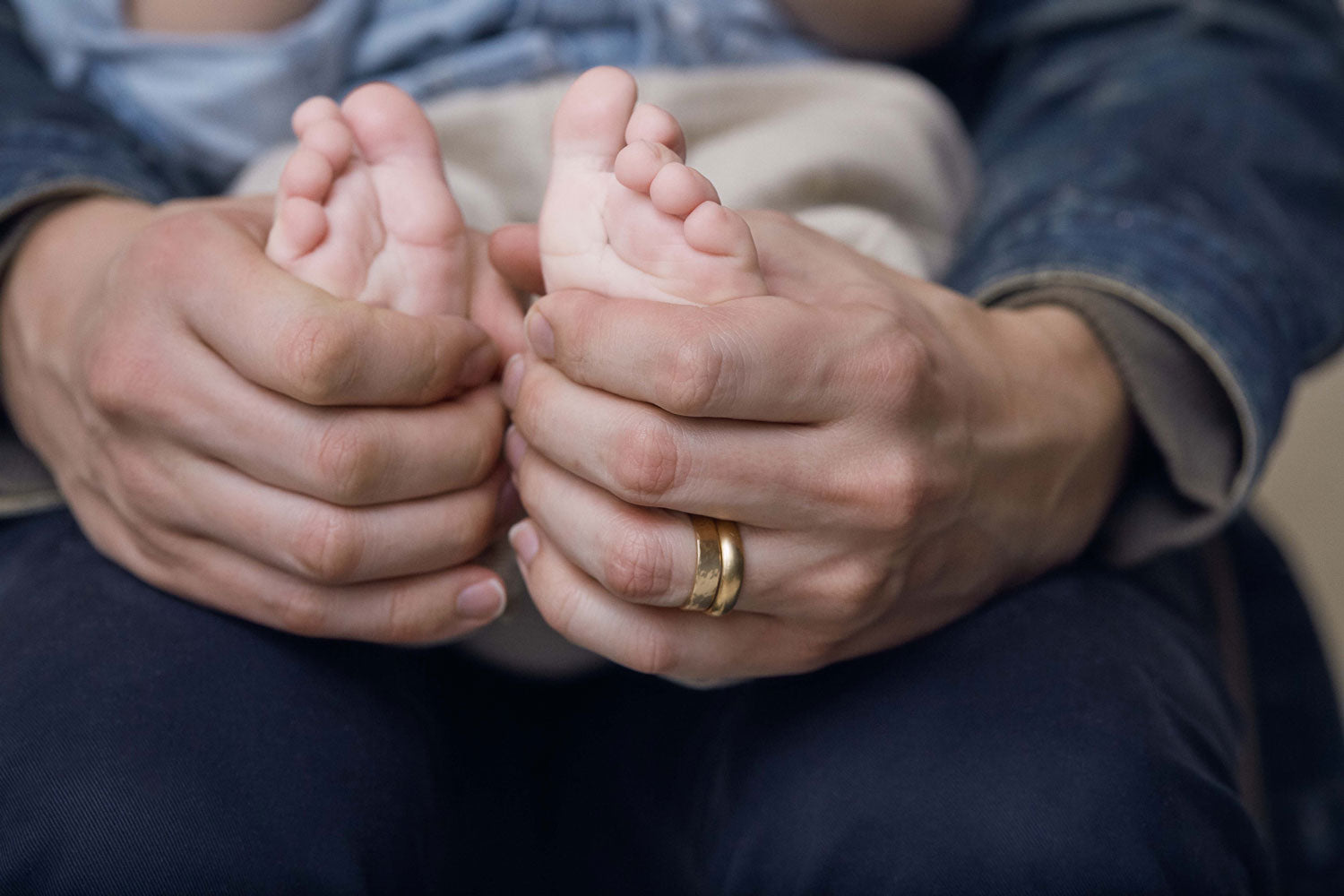 A baby sit's on their father's lap. The father's hands are gently wrapped around the baby toes while the father wears a men's wedding band by Anna Sheffield.