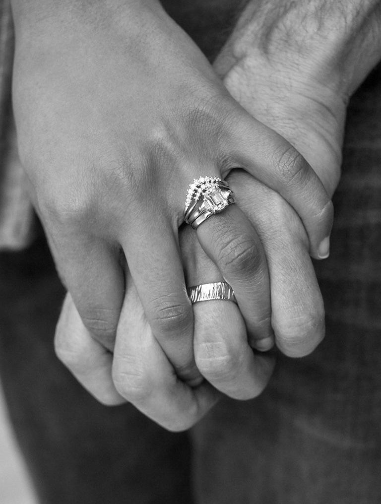 A black and white image of 2 holding hands, one hand is wearing a three stone engagement ring with matching curved wedding bands, and the other hand is wearing a textured classic men's wedding band.