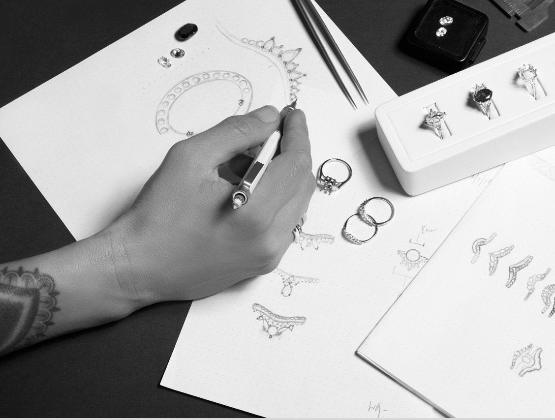 An image of hand with tattoos on the forearm holds a pencil above a paper with ring sketches during a custom consultation. There are scattered diamonds and ring settings on top of the sketches. 