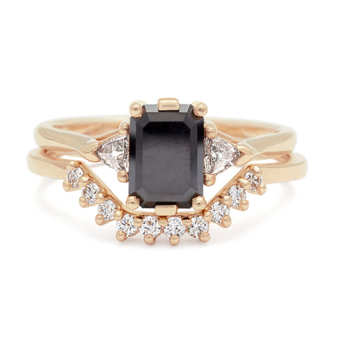 A three stone engagement ring with an emerald cut and emerald shape 7x5mm black diamond center stone with two white diamond trillion side stones set in yellow gold set above a white diamond nesting band. 5