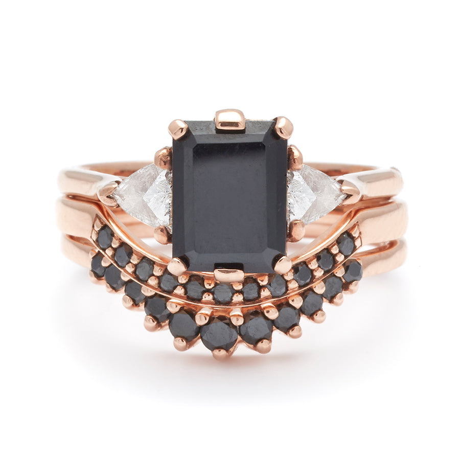 A three stone engagement ring with an emerald cut and emerald shape black diamond center stone with two white diamond trillion side stones set in rose gold above two black diamond curve bands.