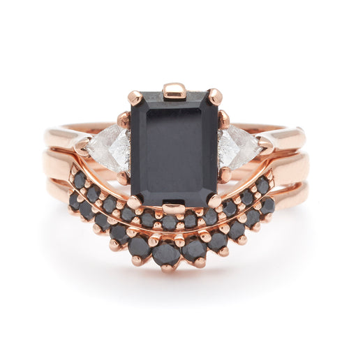 A three stone engagement ring with an emerald cut and emerald shape black diamond center stone with two white diamond trillion side stones set in rose gold above two black diamond curve bands.
