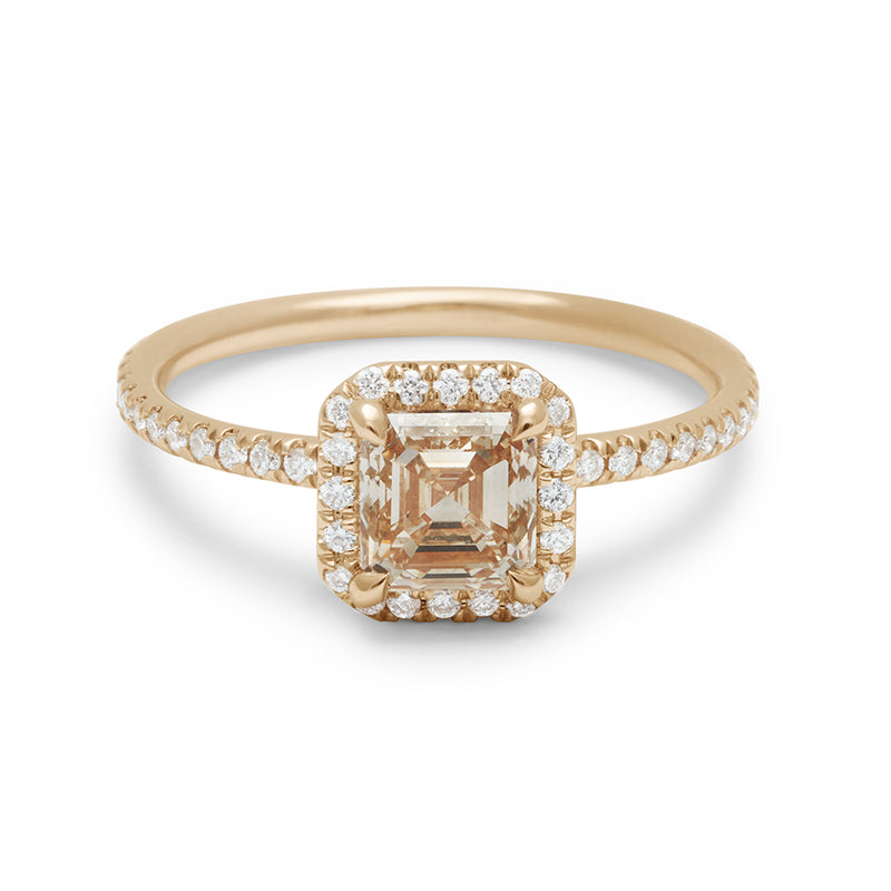 An engagement ring with an asscher shaped champagne diamond center stone surrounded by a white diamond pave halo set in yellow gold with white diamond pave on the band.