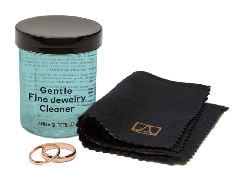 On the left is our gentle fine jewelry cleaner. Two wedding bands are placed below the cleaner and a cloth is set to the right.