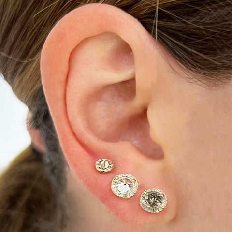 A ear with three diamond halo earrings; two with medium sized center stones and one tiny center stone. All set in yellow gold with stones black rutilated quartz, white diamond, and champagne diamond (from bottom to top)., White Gold, Rose Gold, Yellow Gold 5