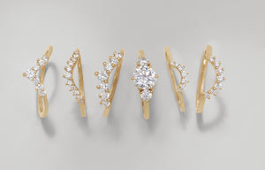 alt="Five white diamond curve bands lined up with one three stone white diamond engagement ring on a grey and white background."