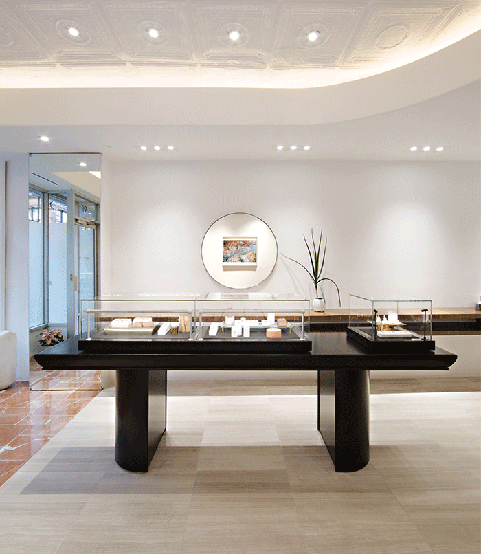 An airy and bright NYC jewelry showroom, with a large jewelry case in the center. There are soothing, bright lights with plants and a rounded mirror hung on the wall above the jewelry case.