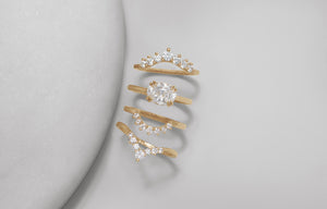 alt="Three white diamond curve bands lined up with one east/west set solitaire white diamond engagement ring next to a grey circular stone on a grey and white background."