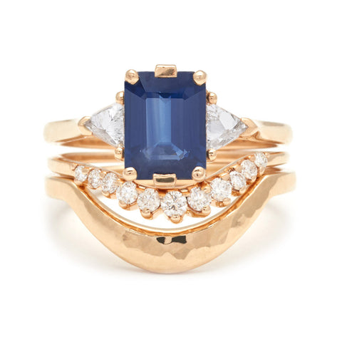A three stone engagement ring with a blue sapphire center stone stacked above a white diamond nesting band and a solid gold hammered nesting band all set in yellow gold.