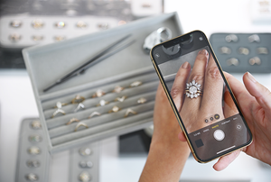 A light-complexion hand holding a phone looking at a white diamond engagement ring stack. Set in front of many jewelry trays with an assortment of rings and bands.