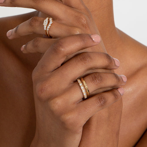 A straight band with leaf like definition set in yellow gold stacked above a white diamond 3mm wheat band on a darker-complextion right hand ring finger. On her left hand holds a five white diamond engagement rings stacked above a 7 white diamond curve band in yellow gold.