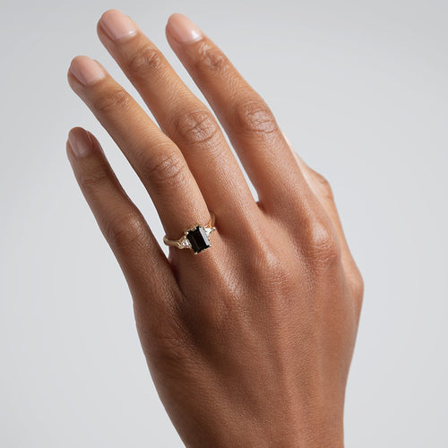 A three stone engagement ring with an emerald cut and emerald shape 8x6mm black diamond center stone with two white diamond trillion side stones set in yellow gold on a medium-complexion hand., White Gold, Rose Gold, Yellow Gold