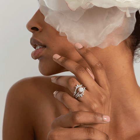 Two asymetrical white diamond scalloped bands with dusted white diamond around the band set in white gold surrounding a solitaire white diamond engagement ring on a darker-complexion ring finger. 4