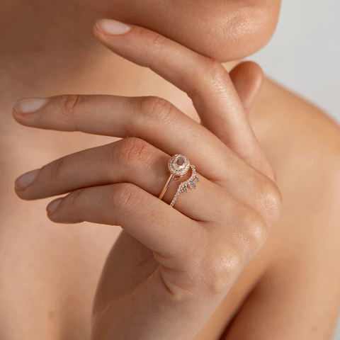 An asymetrical white diamond scalloped band with dusted white diamond around the band set in yellow gold below an oval halo ring on a lighter-complexion ring finger. 5