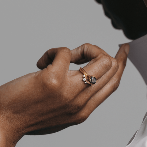 A solid gold band with asymetrical white diamond accent stones set in yellow gold stacked beneath a solitaire east/west grey diamond ring on a dark-complexion ring finger. 6