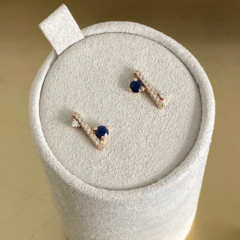 A pair of straight line of white diamond pave with sapphire accent stones on either side of the earrings set in yellow gold on a grey earring box. 5