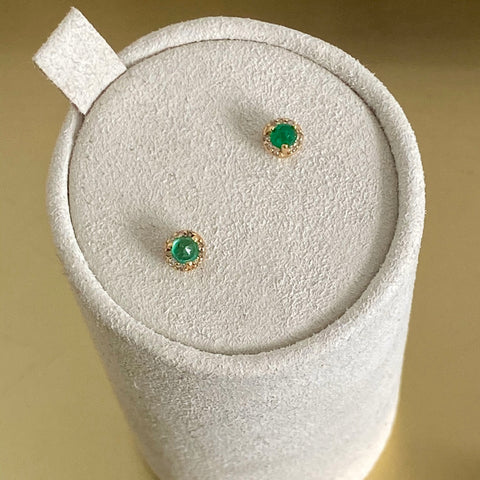 A pair of tiny emerald center stone earrings with a halo of champagne diamond set in yellow gold on a grey earring box. 5