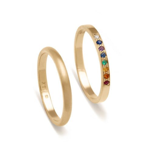 A 2mm classic round band with a brushed finish set in yellow gold next to a 1.5mm band with rainbow gemstones set in yellow gold.