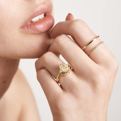 A three stone engagement ring with an emerald shape yellow diamond center stone with two white diamond trillion side stones set in yellow gold above a white diamond curved band on a light-complexion ring finger. 4