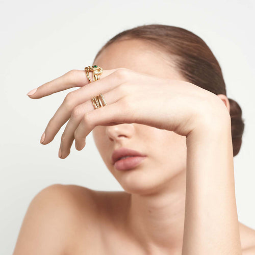 A serpent band with a green emerald center stone and white diamond accent stones around the head of the snake set in yellow gold on a lighter-complextion hand paired with another serpent band.
