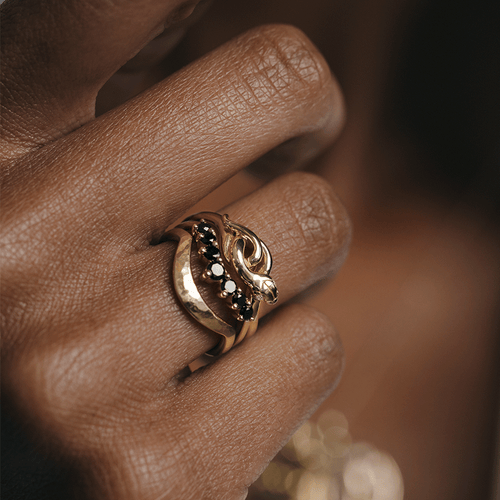 A serpent band with white diamonds for eyes set in yellow gold stacked above a black diamond curve band and a hammered solid gold curve band on a dark-complexion ring finger.