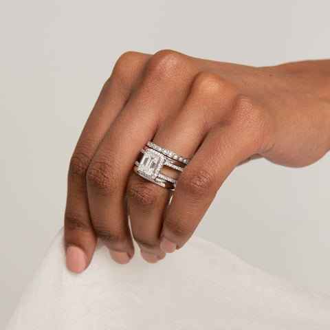 A double banded ring with an emerald cut and shaped white diamond center stone stacked between two solid gold bands set in white gold and one with white diamonds set on a darker-complexion left ring finger. 4