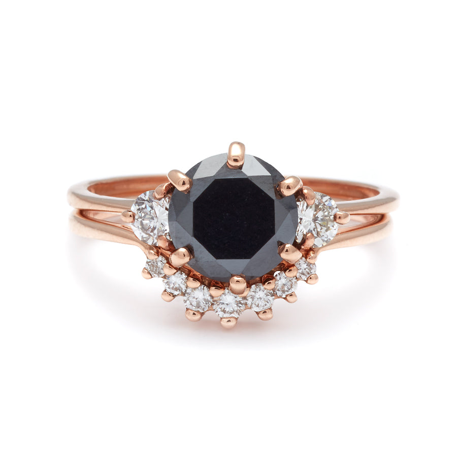 A three stone engagement ring with a round shape black diamond center stone with two white diamond round side stones set in rose gold stacked above a small 7 white diamond curve band in rose gold.