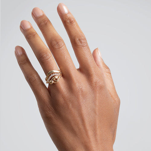A serpent band with white diamonds for eyes set in yellow gold stacked below a white diamond curved band and a solid gold tapered curve band on a medium-complexion hand on a grey background.