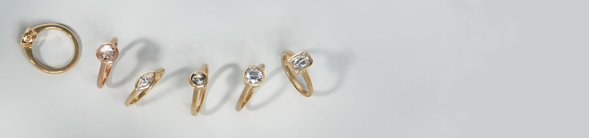 Six engagement rings with bezel set diamonds in round, marquise, and elongated shapes on a grey background.