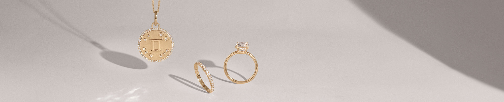 A white diamond halo set zodiac charm on a yellow gold chain with a hidden halo engagement ring next to it and a white diamond eternity band on a grey background.