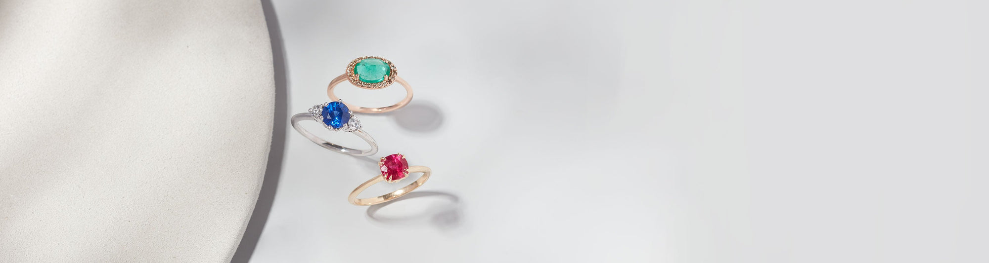 Green emerald, sapphire, and ruby rings stacked next to a circular white stone on a grey background.