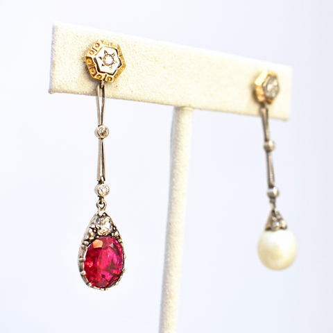Two drop earrings on a white suede stand. A hexagon shaped white gold post featuring a star engraving and encircled in yellow gold drop down into white gold chain links separated by white round diamonds to a round ruby droplet. The earring on the right is the same design except it features a pearl droplet instead of a ruby. 