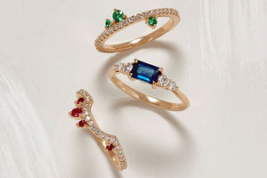 A birds eye view of engagement rings and wedding bands in mixed precious stones on light-grey background with a brushstroke design