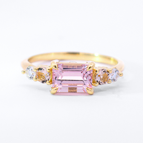 A five stone ring featuring an emerald cut pink morganite center stone and one round morganite and one round white diamond on each side set in yellow gold against a white background. 