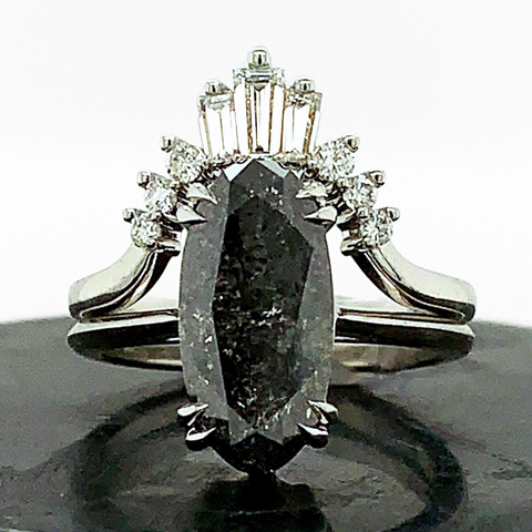 An elongated oval shaped rose cut black diamond ring paired with a nesting band featuring white baguette diamonds and white round diamonds. Both rings are set in platinum and sit on a black riser. 