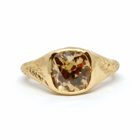 A yellow gold signet ring featuring a cushion cut brown diamond center and leaf engravings on both sides of the band against a white background.