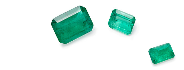 An image of 3 emerald cut green emerald stones, all in different sizes with varying colors of green laying on a flat surface.