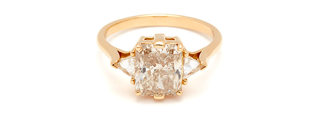 An image of the Bea Three Stone Ring sitting on a flat surface with a champagne diamond radiant cut center and white diamond trillion side stones.