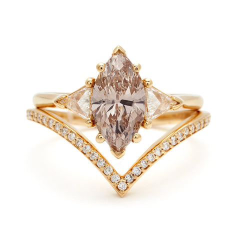 A three stone engagement ring with a marquise shaped champagne diamond center stone with white diamond trillion side stones set in yellow gold stacked on top of a diamond dusted chrysalis band.