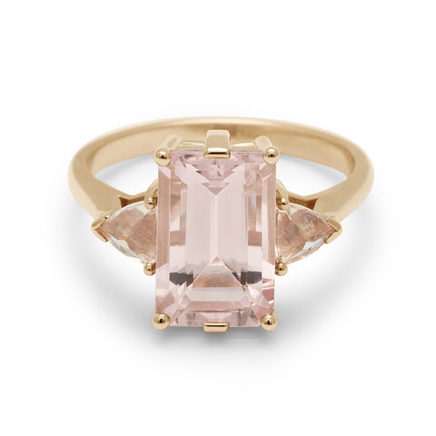 A three stone engagement ring with an emerald shape pink morganite center stone with two peach morganite trillion side stones set in yellow gold.