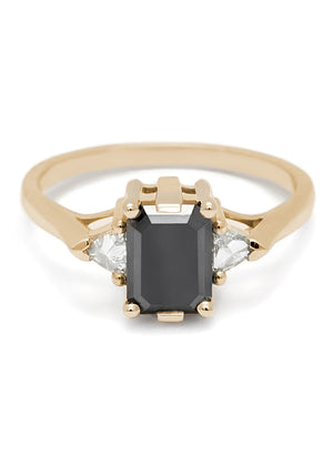 A three stone engagement ring with an emerald cut and emerald shape 7x5mm black diamond center stone with two white diamond trillion side stones set in yellow gold.