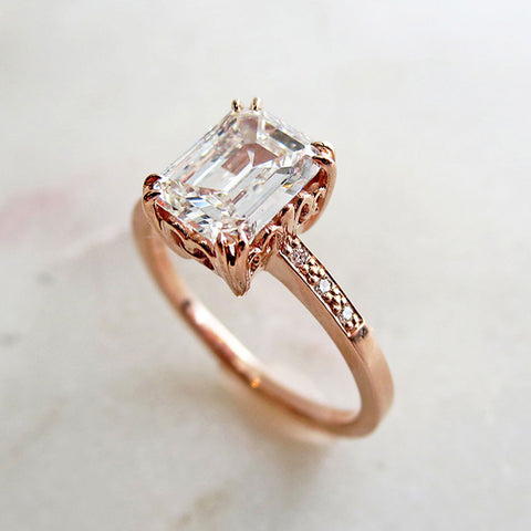 Angled view of a solitaire ring with an emerald shape and cut champagne diamond with white diamond pave on either side set in rose gold on a pink background.