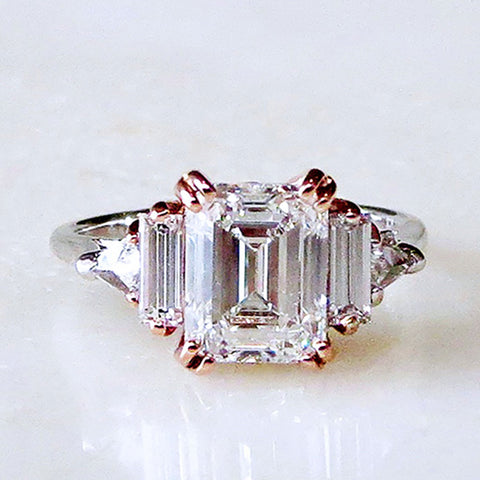 A ring with an emerald cut and shaped white diamond center stone with one baguette white diamond and one white diamond trillion on either side with rose gold claw prongs set in white gold on a grey background.