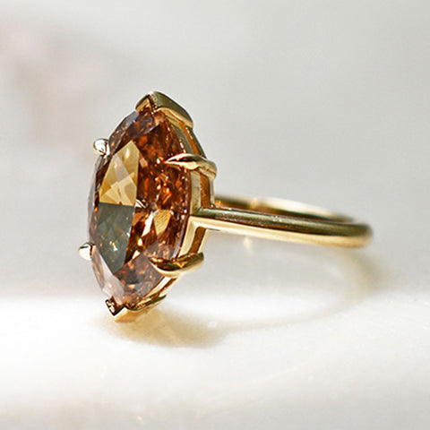 An solitaire ring with a marquise shape champagne diamond stone set in a yellow gold band on a grey background.