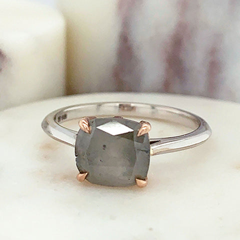 An east/west solitaire ring with an oval shape grey diamond stone set in a rose gold basket and a white gold band on a marble stone.