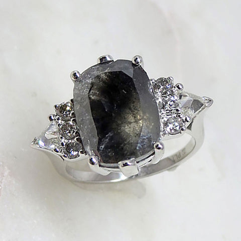 A emerald shape grey diamond center stone ring with three round white diamonds on either and two white diamond trillions on the outer edges of each side set in white gold on a grey background.