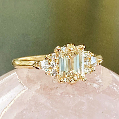 A emerald shape champagne diamond center stone ring with three round white diamonds on either and two white diamond trillions on the outer edges of each side set in yellow gold on a pink stone.
