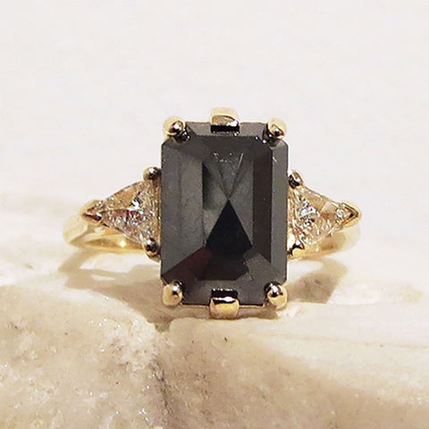 A three stone engagement ring with an emerald shape black diamond center stone with two white diamond trillion side stones set in yellow gold on a white rock.