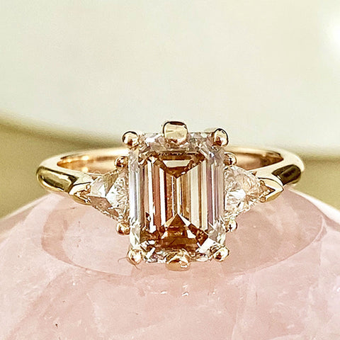 A three stone engagement ring with an emerald cut and emerald shape champagne diamond center stone with two white diamond trillion side stones set in yellow gold.